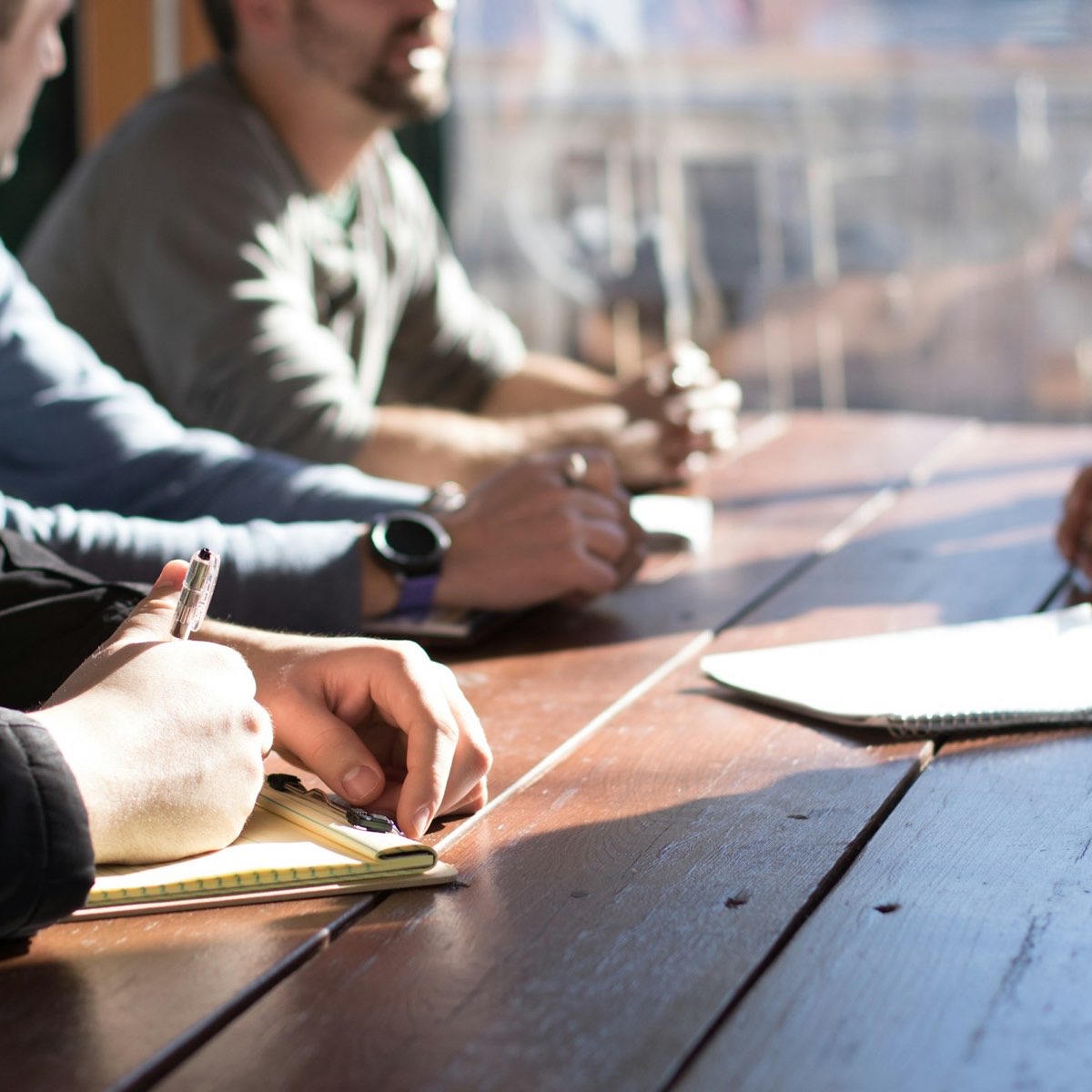 Colleagues seated along a wooden table in a bright room, one taking notes with a pen during a review.