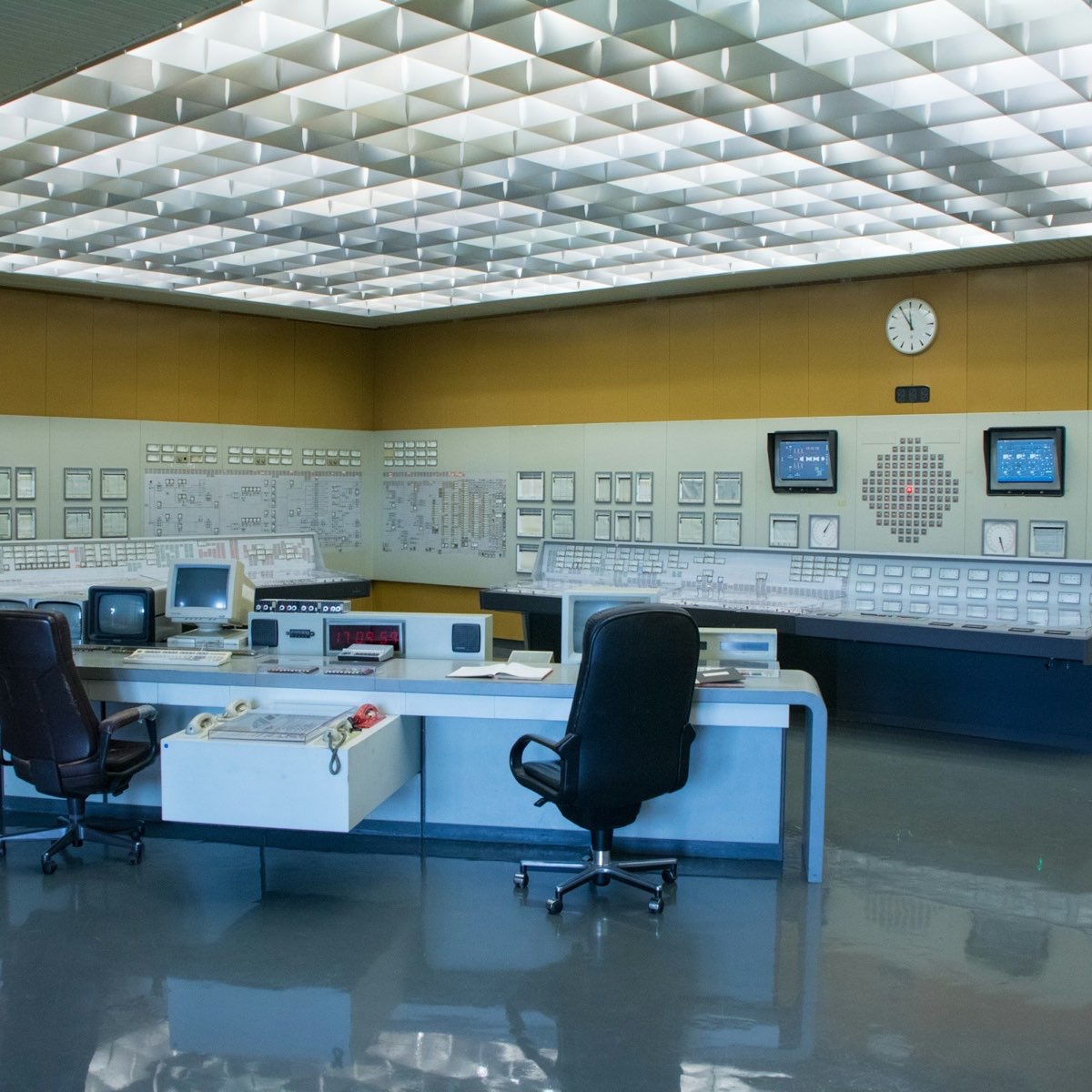 A large industrial control room with long instrument panels on the walls and two empty chairs at a central desk.