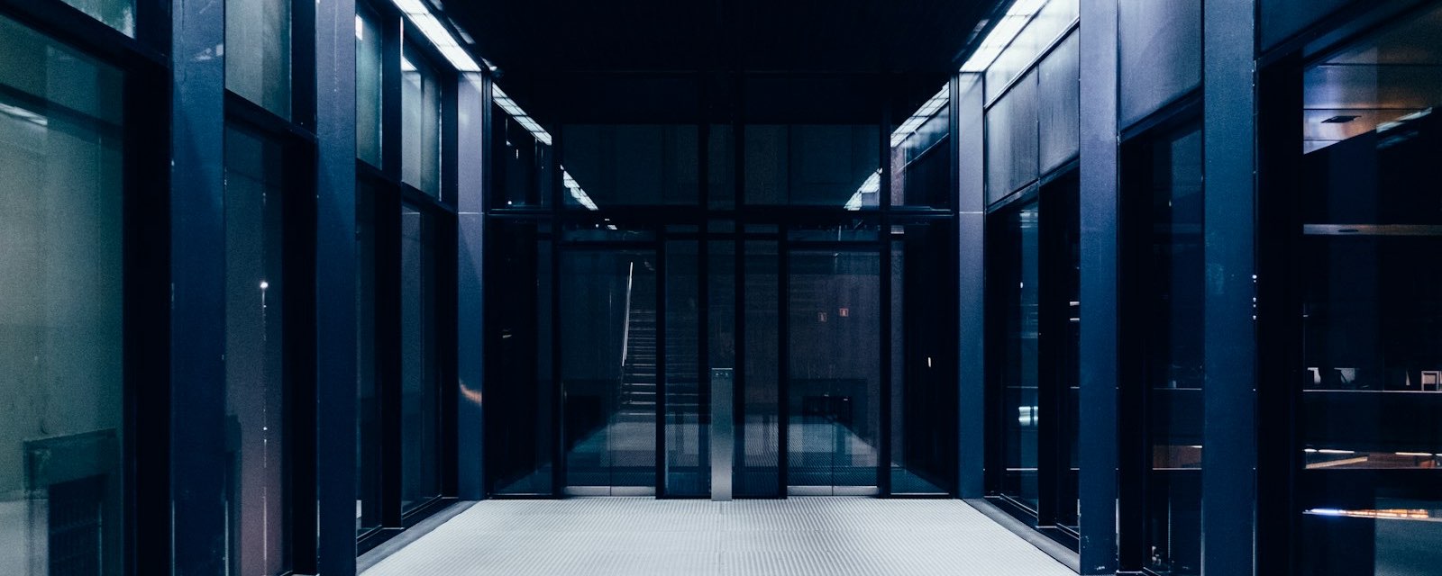A long, dimly lit corridor lined with dark glass panels and doors, ending in a locked glass entrance.