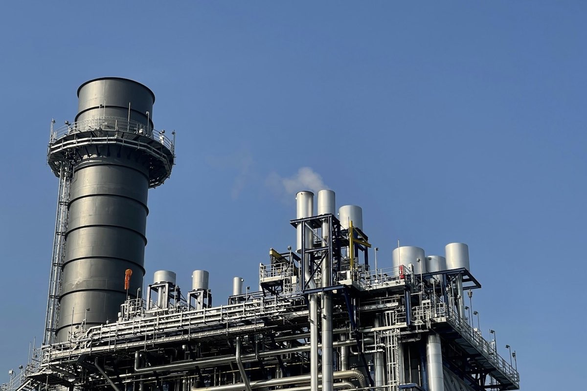 A gas processing plant with a tall steel stack and pipework against a clear blue sky.