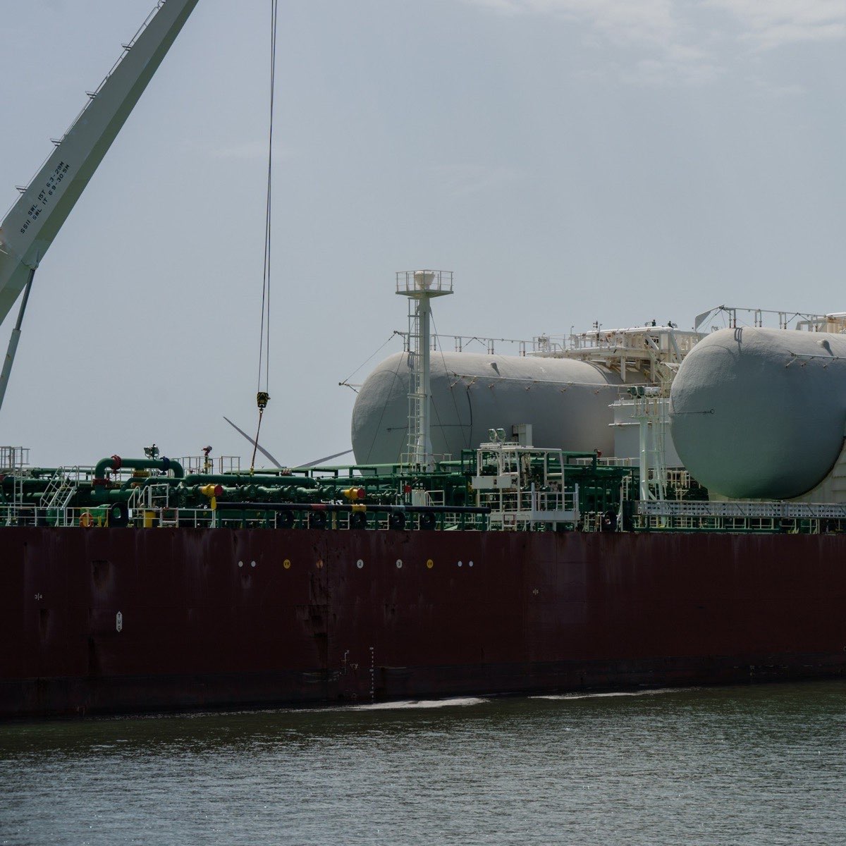 A gas carrier with large cylindrical cargo tanks and green deck pipework, moored in a harbour under a grey sky.