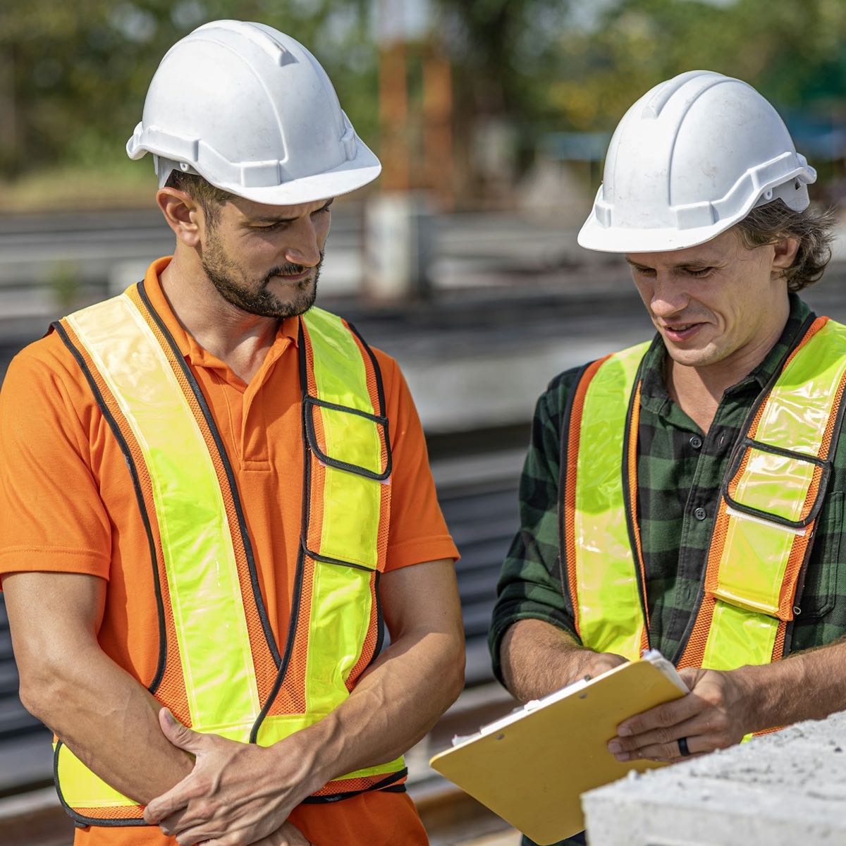 Two engineers in white hard hats and high-visibility vests read a clipboard together on an outdoor work site.