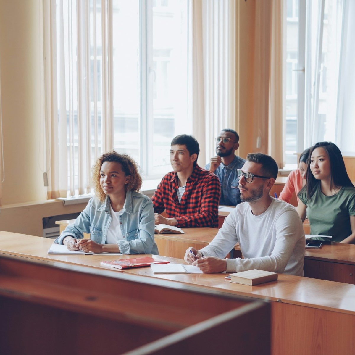 A lecturer talks to students seated at desks in a sunlit classroom.